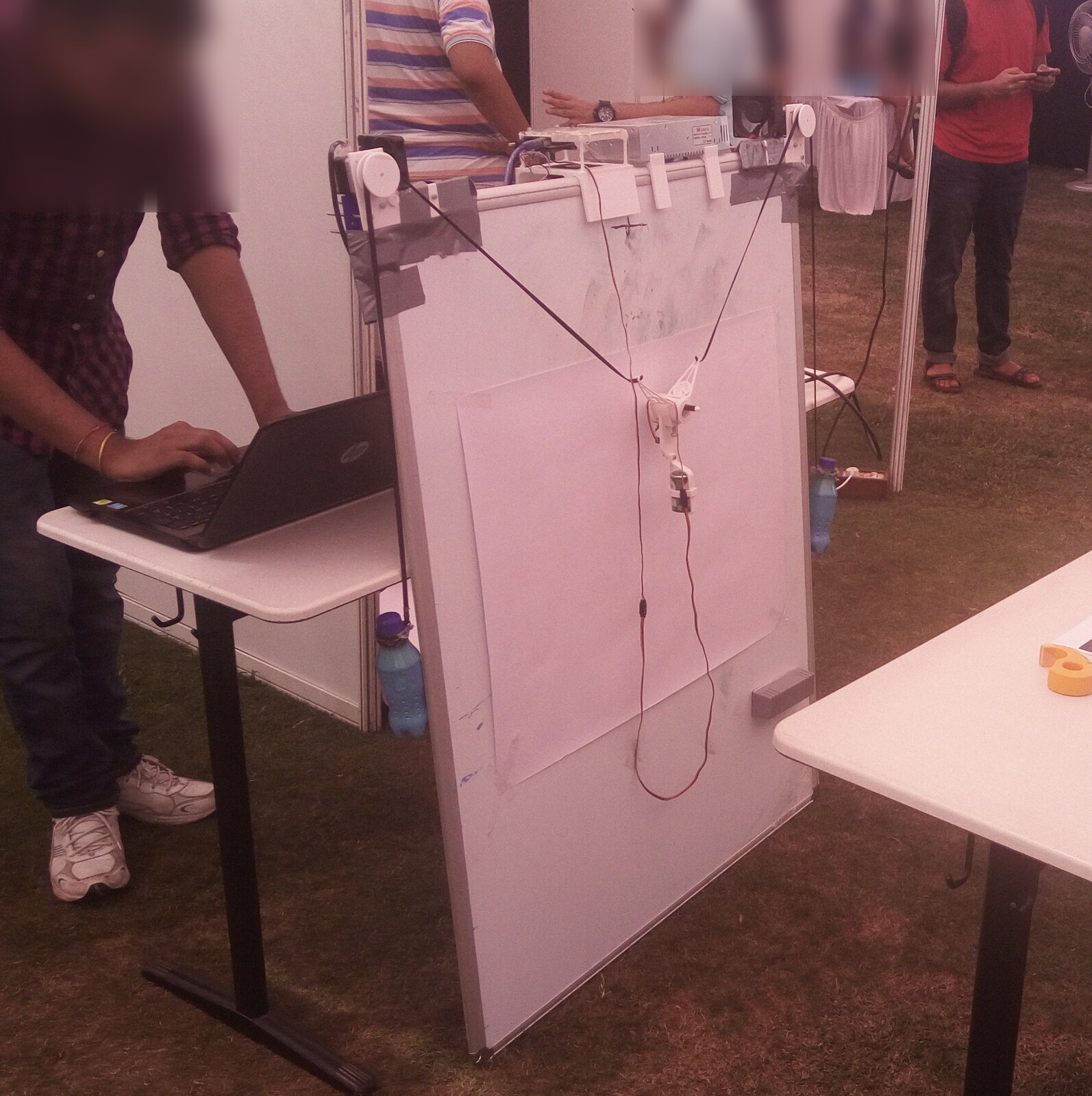 Bono at the Quark booth on the library lawns: whiteboard on a stand, cords running from corner pulleys to the pen gondola, the laptop driving it