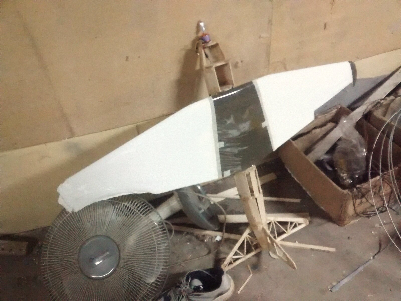 A built-up airframe leaning against the workshop wall: white molded wing, balsa truss fuselage, motor on a wooden pylon