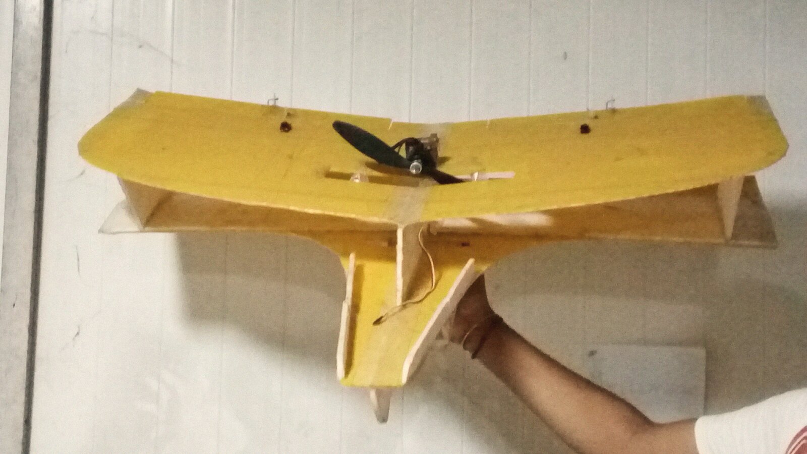 A yellow swept flying wing held up for inspection, with tip fins and the motor in a mid-wing cutout