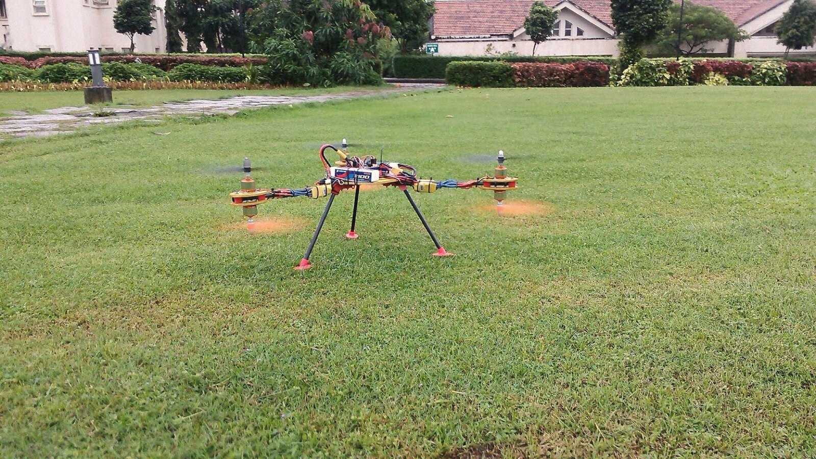 The quadcopter hovering low over a campus lawn, propellers blurred, football practice continuing in the background