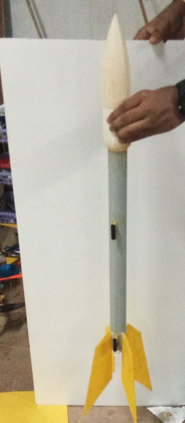 The model rocket held up for inspection: shaped nose cone, pipe body, yellow foam fins