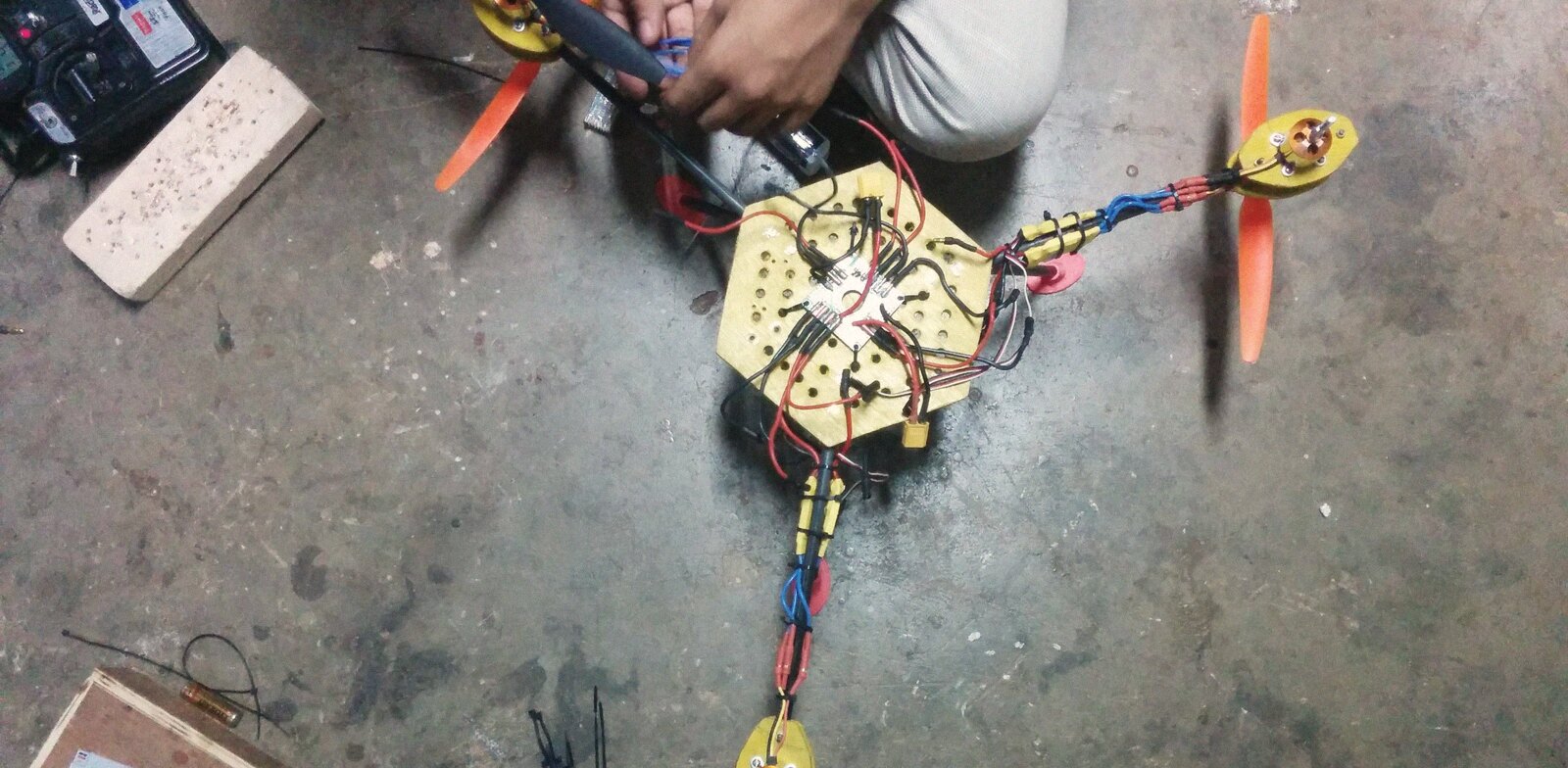 A Y-frame tricopter on the workshop floor mid-build: plywood center plate, power distribution wiring half soldered, the transmitter waiting in the corner