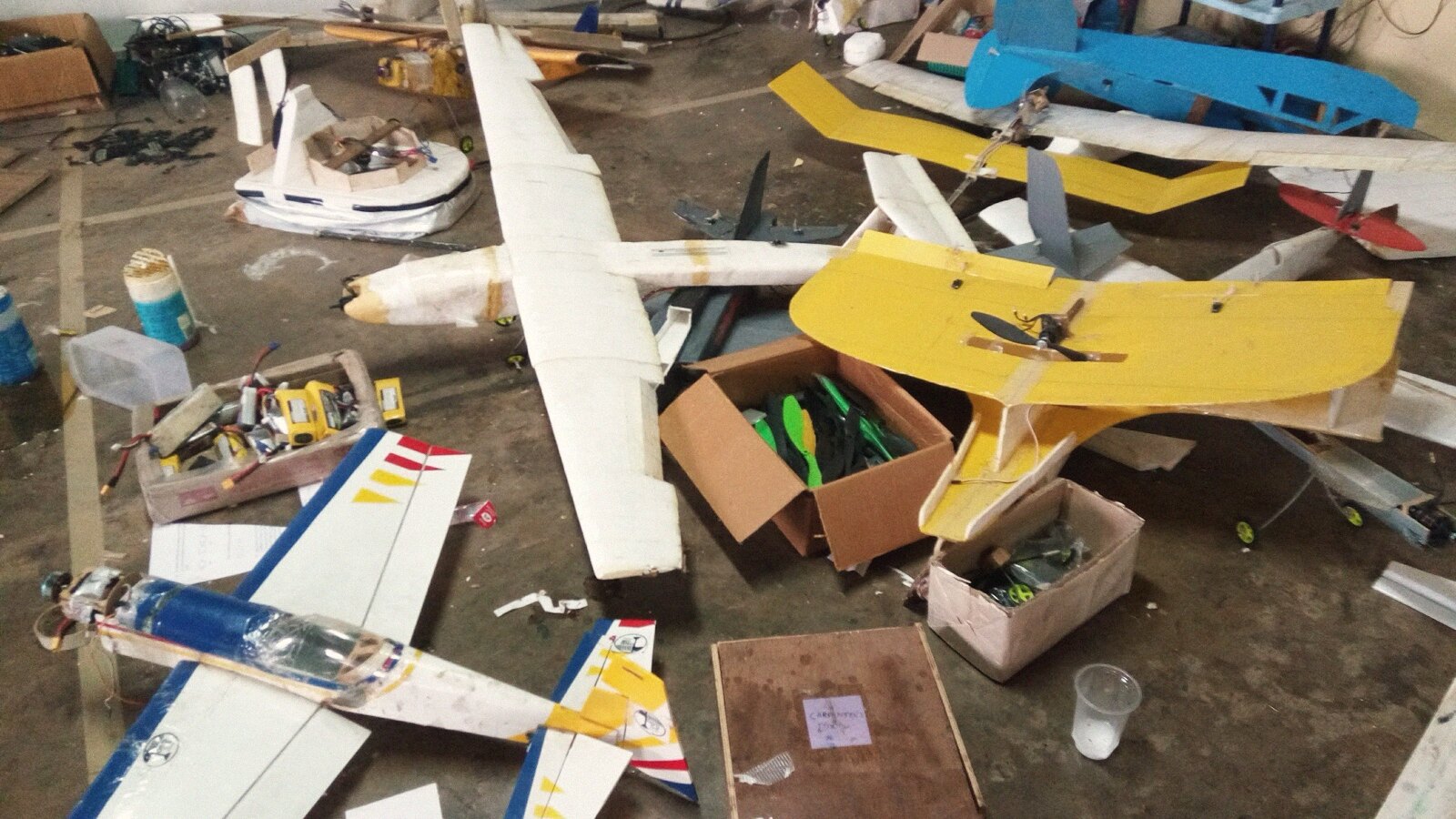The workshop floor: a long-winged white plane, a yellow trainer, a blue balsa build, boxes of propellers and LiPo packs, and a hovercraft where furniture should be