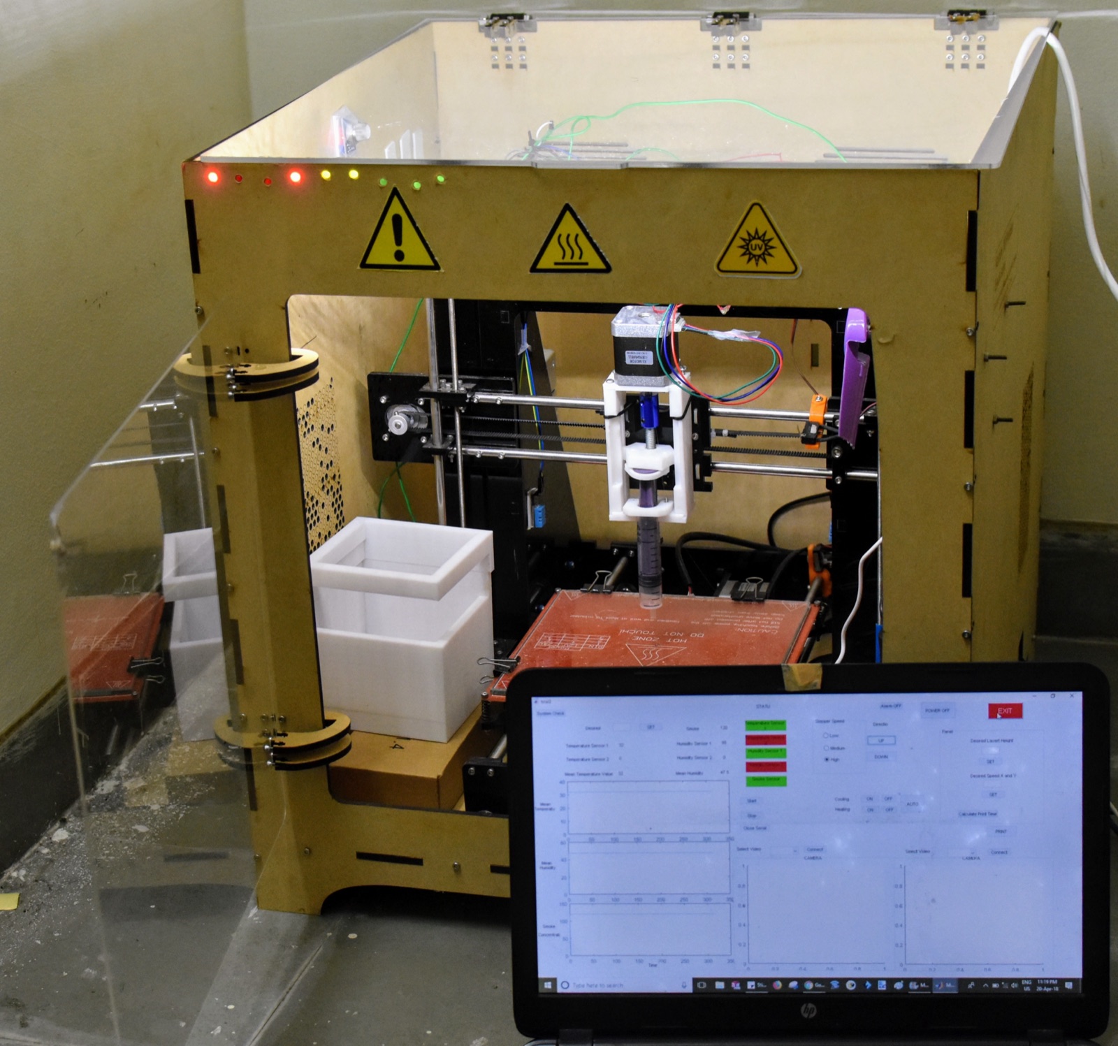 The BioP printer: laser-cut enclosure with UV and hot-surface warnings, syringe extruder over the heated bed, and the control software running in front