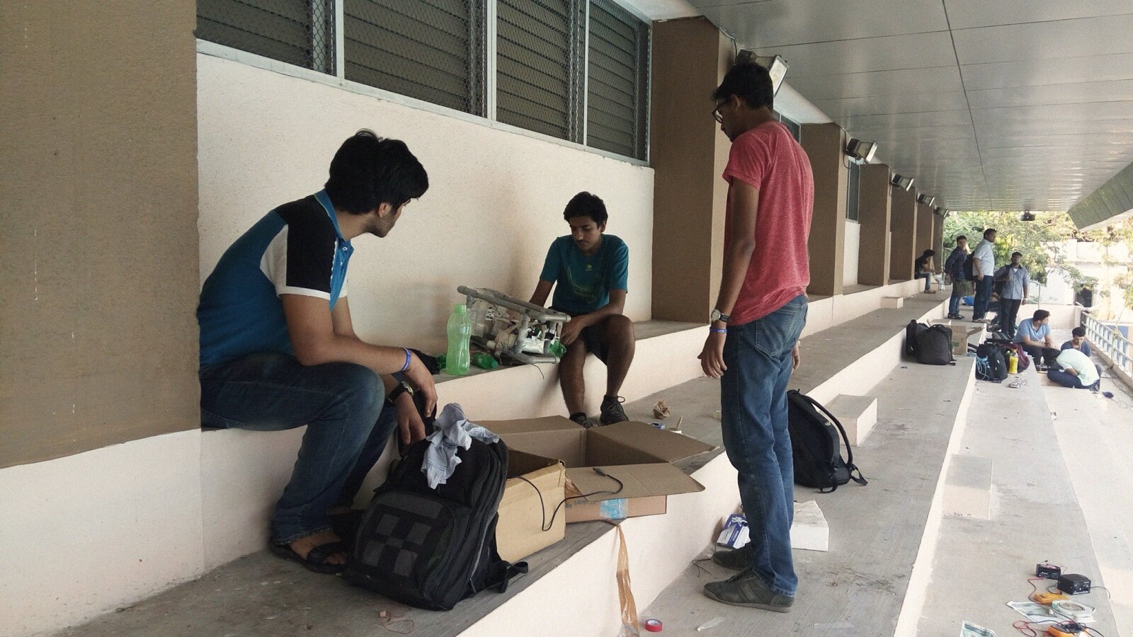 The frame out for a December test day: teammates on a corridor bench, cardboard boxes, chargers and a multimeter spread down the steps